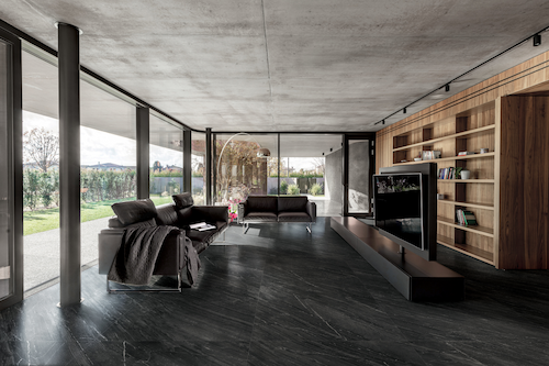 Nowoczesny salon z podłogą z płytek Florim Biotech Soap Stone w ciemnym, kamiennym wykończeniu, duże przeszklenia i zabudowa z drewna przy ścianie TV.